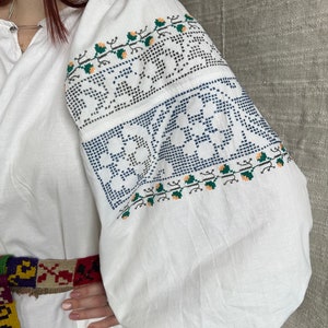 May include: White embroidered blouse with a floral pattern in blue, green, and orange. The blouse has long sleeves and a gathered neckline.