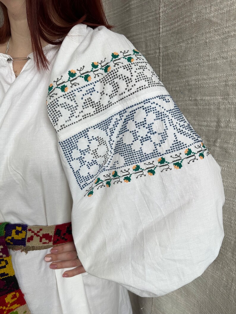 May include: White embroidered blouse with a floral pattern in blue, green, and orange. The blouse has long sleeves and a gathered neckline.