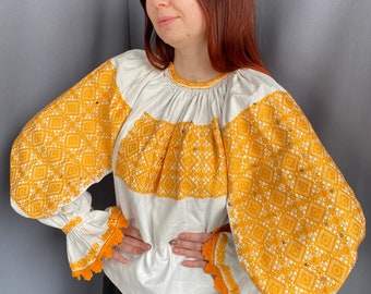 May include: A white blouse with long sleeves and a yellow embroidered pattern. The sleeves have a gathered look and a crocheted trim.