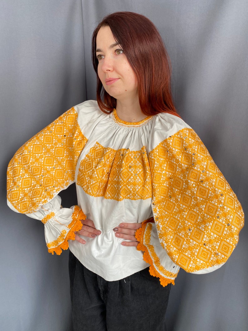 May include: A white blouse with long sleeves and a yellow embroidered pattern. The sleeves have a gathered look and a crocheted trim.
