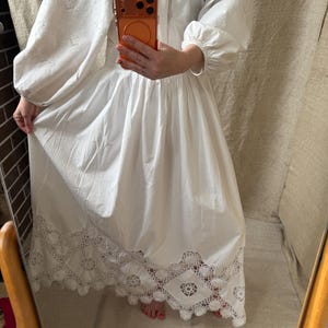May include: A white dress with long, puffy sleeves and a full skirt. The sleeves and upper bodice feature intricate embroidered cutwork. The hemline is detailed with a delicate, openwork lace trim. The dress is made of a lightweight fabric.