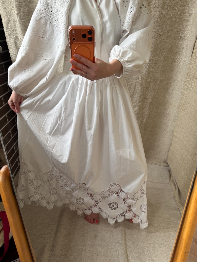 May include: A white dress with long, puffy sleeves and a full skirt. The sleeves and upper bodice feature intricate embroidered cutwork. The hemline is detailed with a delicate, openwork lace trim. The dress is made of a lightweight fabric.