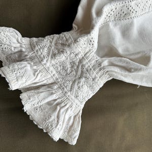 Op de afbeelding: Witte katoenen stof met een delicate kantversiering. De stof is geplooid en gerimpeld aan de rand, wat een zachte en vrouwelijke uitstraling cre&euml;ert.