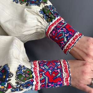 May include: White linen shirt with colorful embroidered details on the sleeves and cuffs. The embroidery features geometric patterns in red, blue, green, and black.