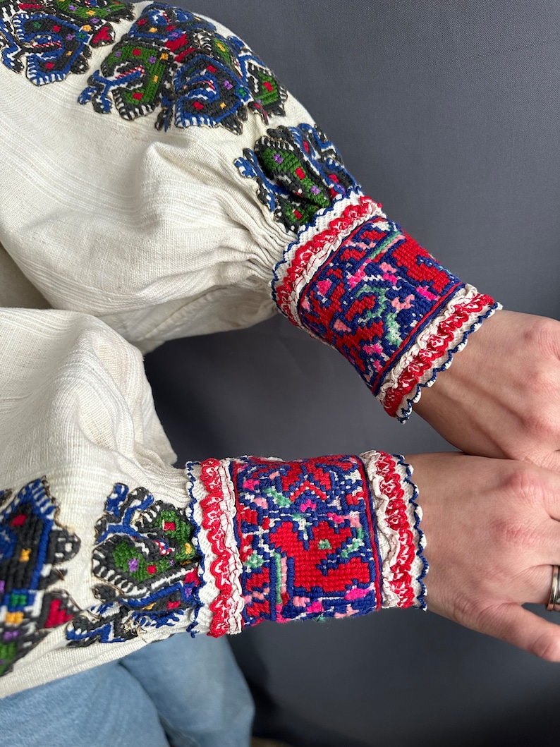 May include: White linen shirt with colorful embroidered details on the sleeves and cuffs. The embroidery features geometric patterns in red, blue, green, and black.