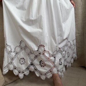 May include: A white dress with a detailed, openwork lace hem. The dress has long sleeves and a full skirt. The hem features a repeating pattern of geometric shapes and circular cutouts. The fabric appears to be cotton or a similar lightweight material.
