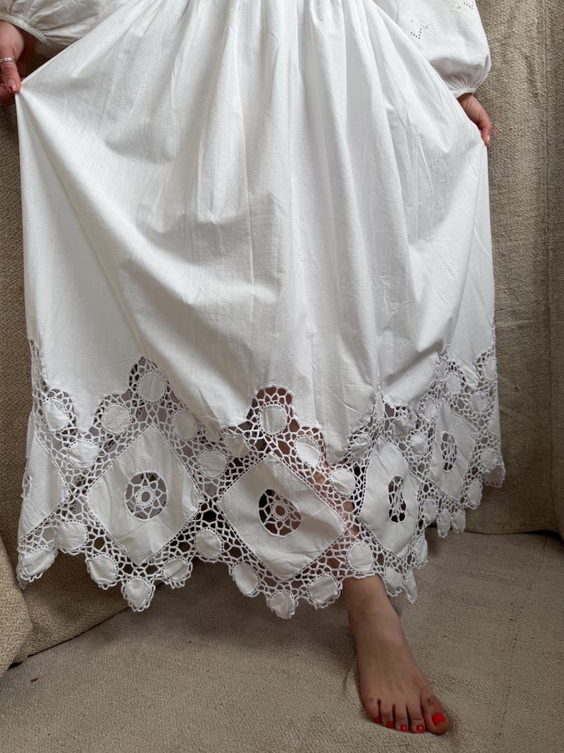 May include: A white dress with a detailed, openwork lace hem. The dress has long sleeves and a full skirt. The hem features a repeating pattern of geometric shapes and circular cutouts. The fabric appears to be cotton or a similar lightweight material.