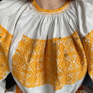 May include: White embroidered blouse with a yellow geometric pattern and sequins. The blouse has a gathered neckline and long sleeves with a crocheted trim.