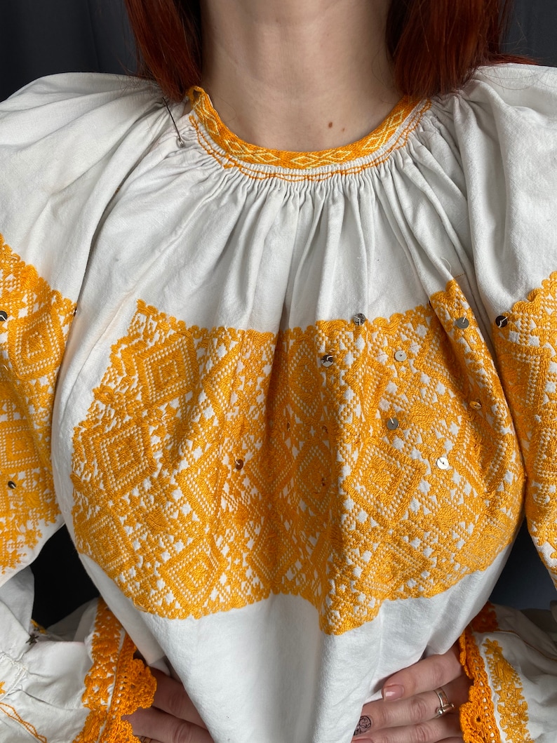 May include: White embroidered blouse with a yellow geometric pattern and sequins. The blouse has a gathered neckline and long sleeves with a crocheted trim.