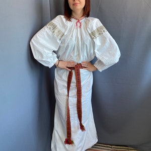 May include: A white embroidered blouse with long sleeves and a gathered neckline. The blouse is worn with a white skirt and a brown woven belt with colorful accents.