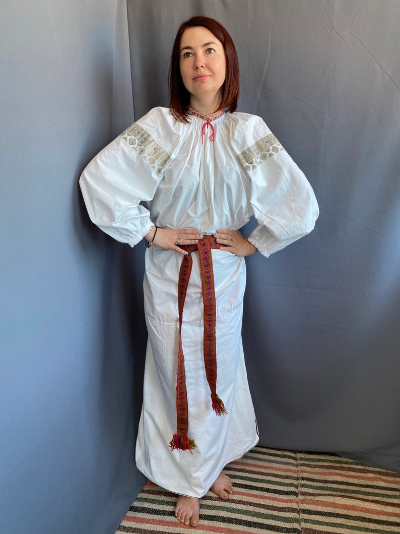 May include: A white embroidered blouse with long sleeves and a gathered neckline. The blouse is worn with a white skirt and a brown woven belt with colorful accents.