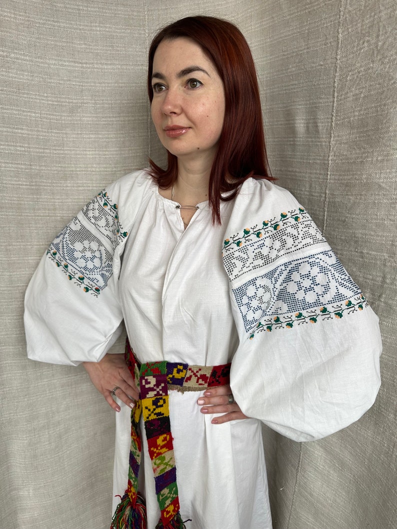 May include: A white embroidered blouse with long sleeves and a colorful woven belt. The blouse has a traditional Ukrainian design with floral patterns.