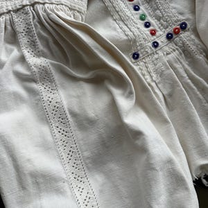 Op de afbeelding: Wit linnen blouse met een kantversiering en gekleurde knopen. De blouse heeft een geplooide halslijn en lange mouwen.