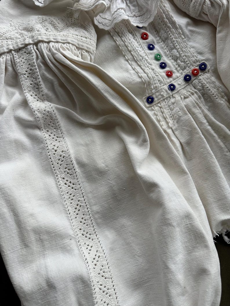 Op de afbeelding: Wit linnen blouse met een kantversiering en gekleurde knopen. De blouse heeft een geplooide halslijn en lange mouwen.