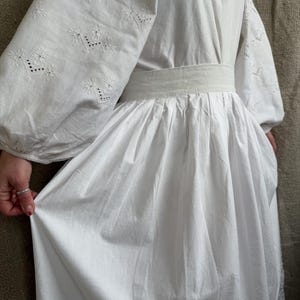 May include: A white dress with long, puffy sleeves and intricate embroidered floral patterns. The dress features a cinched waist and a flowing skirt. The sleeves have detailed embroidery near the cuffs. The dress is made of a lightweight fabric.