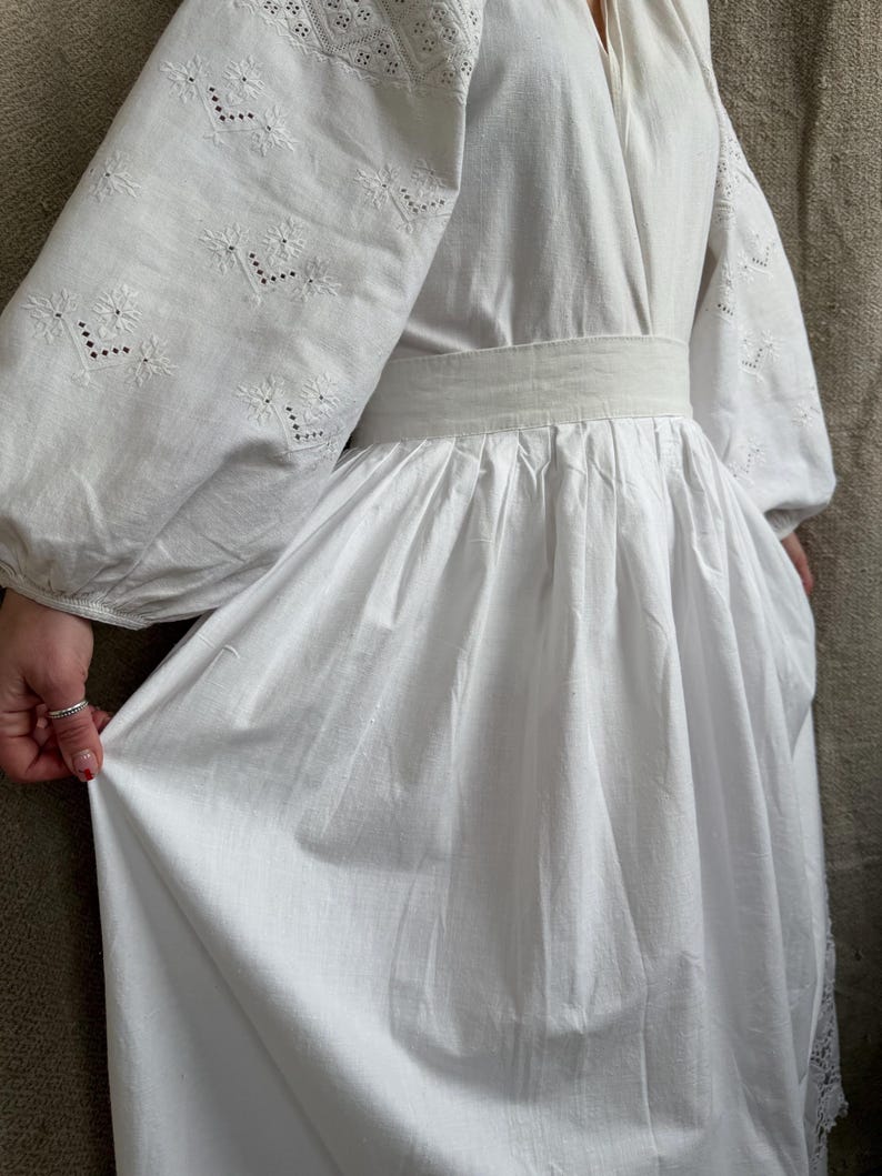 May include: A white dress with long, puffy sleeves and intricate embroidered floral patterns. The dress features a cinched waist and a flowing skirt. The sleeves have detailed embroidery near the cuffs. The dress is made of a lightweight fabric.