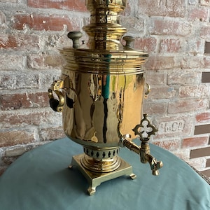 May include: A traditional brass samovar with a decorative spout and handles. The samovar is on a square base and has a polished finish.