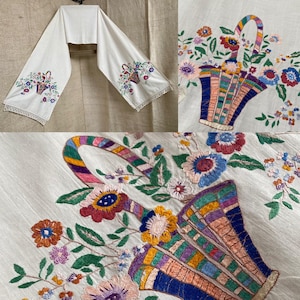 May include: White embroidered table runner with colorful floral basket design. The embroidery features a variety of flowers in shades of red, blue, purple, and yellow. The basket has a striped handle and is filled with flowers.