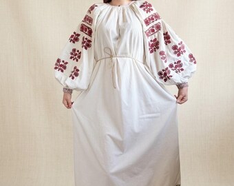May include: A long white dress featuring voluminous sleeves with dark red floral embroidery. The garment includes a gathered neckline, a thin waist tie, and a decorative dark grey patterned border along the hemline. The fabric is plain white.
