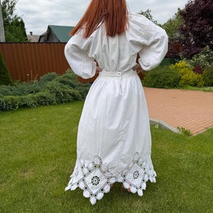 May include: A white, long-sleeved dress with intricate embroidery on the sleeves and a detailed, openwork lace hem. The dress features a fitted waist and a full skirt, creating a flowing silhouette. The person is standing on green grass.