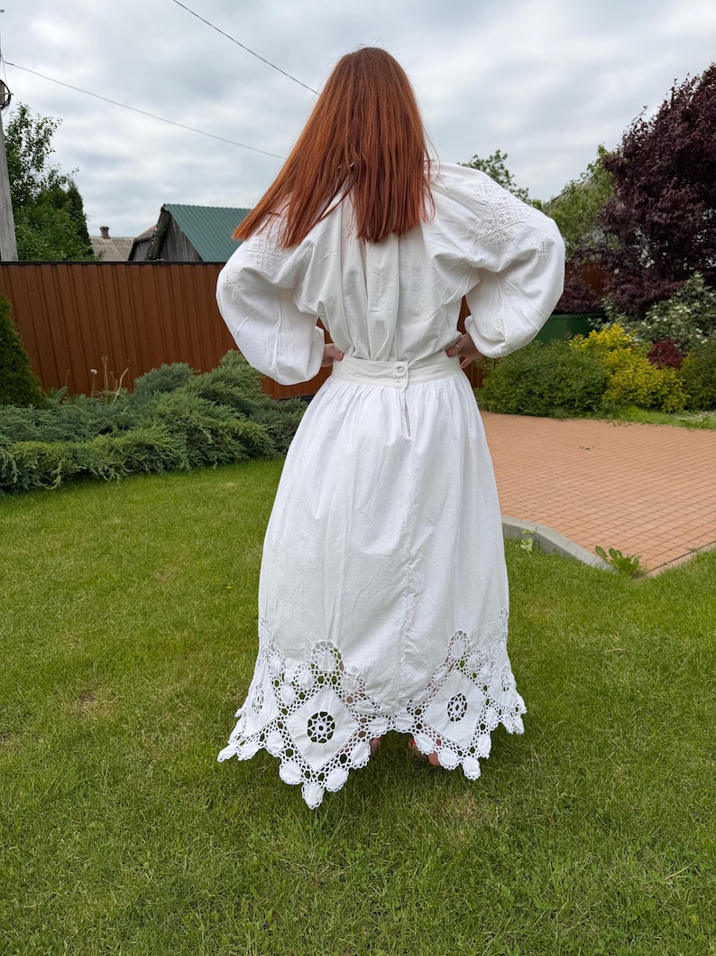 May include: A white, long-sleeved dress with intricate embroidery on the sleeves and a detailed, openwork lace hem. The dress features a fitted waist and a full skirt, creating a flowing silhouette. The person is standing on green grass.