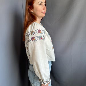 May include: A white linen blouse with embroidered floral details in shades of red, blue, and yellow. The blouse has long sleeves and a slightly cropped fit.