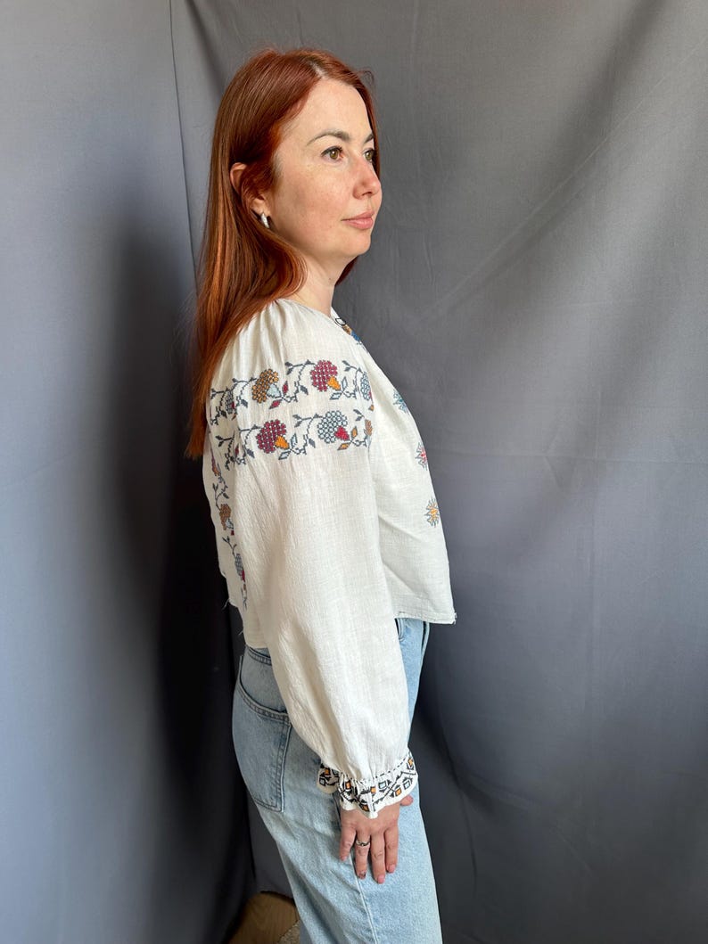 May include: A white linen blouse with embroidered floral details in shades of red, blue, and yellow. The blouse has long sleeves and a slightly cropped fit.