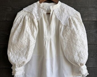 May include: An off-white blouse featuring voluminous puff sleeves with intricate eyelet embroidery, a delicate lace collar, and a button-down placket. The garment has gathered cuffs finished with lace trim and a relaxed, flowing silhouette.