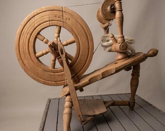 May include: A light brown wooden spinning wheel featuring a large circular wheel with spokes, a flyer assembly with a bobbin, a treadle foot pedal, and a bundle of raw wool fiber resting on the horizontal frame supported by four turned legs.
