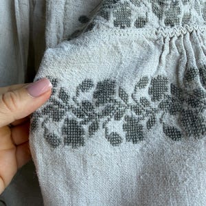 May include: A beige linen blouse featuring intricate dark grey cross-stitch embroidery in a floral pattern along the neckline and shoulder area, with gathered fabric details and a drawstring tie at the collar.
