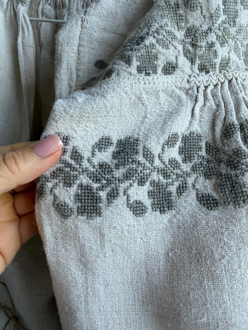 May include: A beige linen blouse featuring intricate dark grey cross-stitch embroidery in a floral pattern along the neckline and shoulder area, with gathered fabric details and a drawstring tie at the collar.