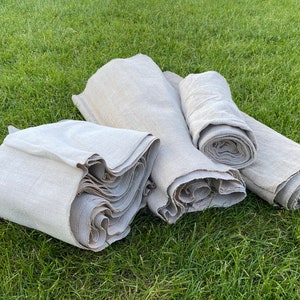 May include: Several rolls and folded stacks of natural linen fabric are arranged on a bed of green grass. The fabric has a light, neutral color and a slightly textured appearance. The edges of the fabric are visible, showing the layers.