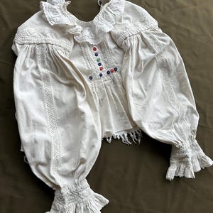Op de afbeelding: Een wit katoenen blouse met een gerimpelde kraag en manchetten. De blouse heeft een decoratief geborduurd ontwerp aan de voorkant en is versierd met gekleurde knopen.
