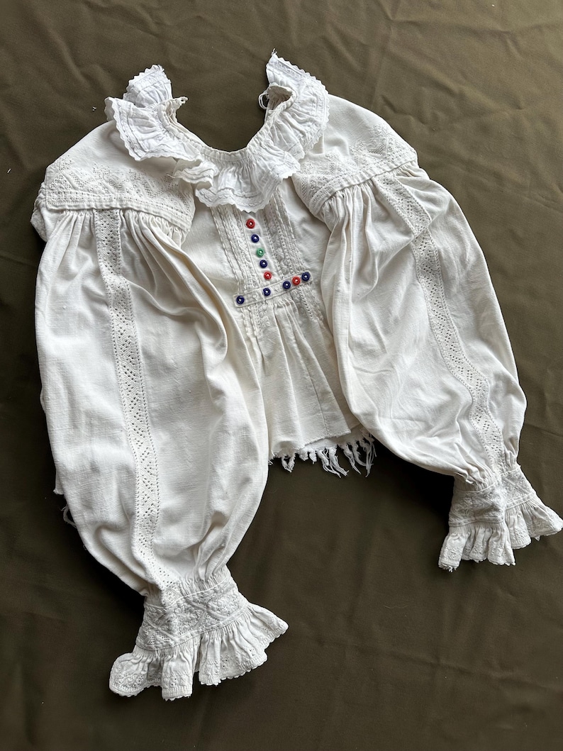 Op de afbeelding: Een wit katoenen blouse met een gerimpelde kraag en manchetten. De blouse heeft een decoratief geborduurd ontwerp aan de voorkant en is versierd met gekleurde knopen.