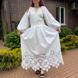 May include: A white, long-sleeved dress with intricate embroidery on the sleeves and a detailed lace hem. The dress features a cinched waist and a flowing skirt. The person is wearing black sandals.