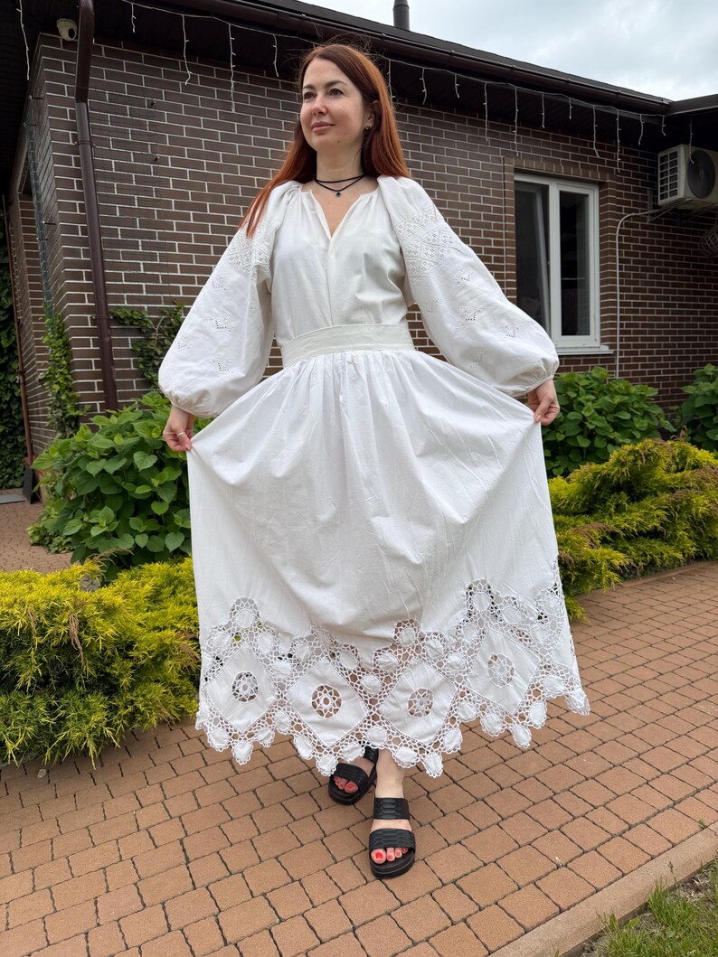 May include: A white, long-sleeved dress with intricate embroidery on the sleeves and a detailed lace hem. The dress features a cinched waist and a flowing skirt. The person is wearing black sandals.