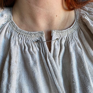 May include: A beige, textured linen blouse featuring a round neckline with gathered fabric details and a vertical slit opening at the centre. The garment is fastened at the top of the opening with a simple silver-toned metal safety pin.