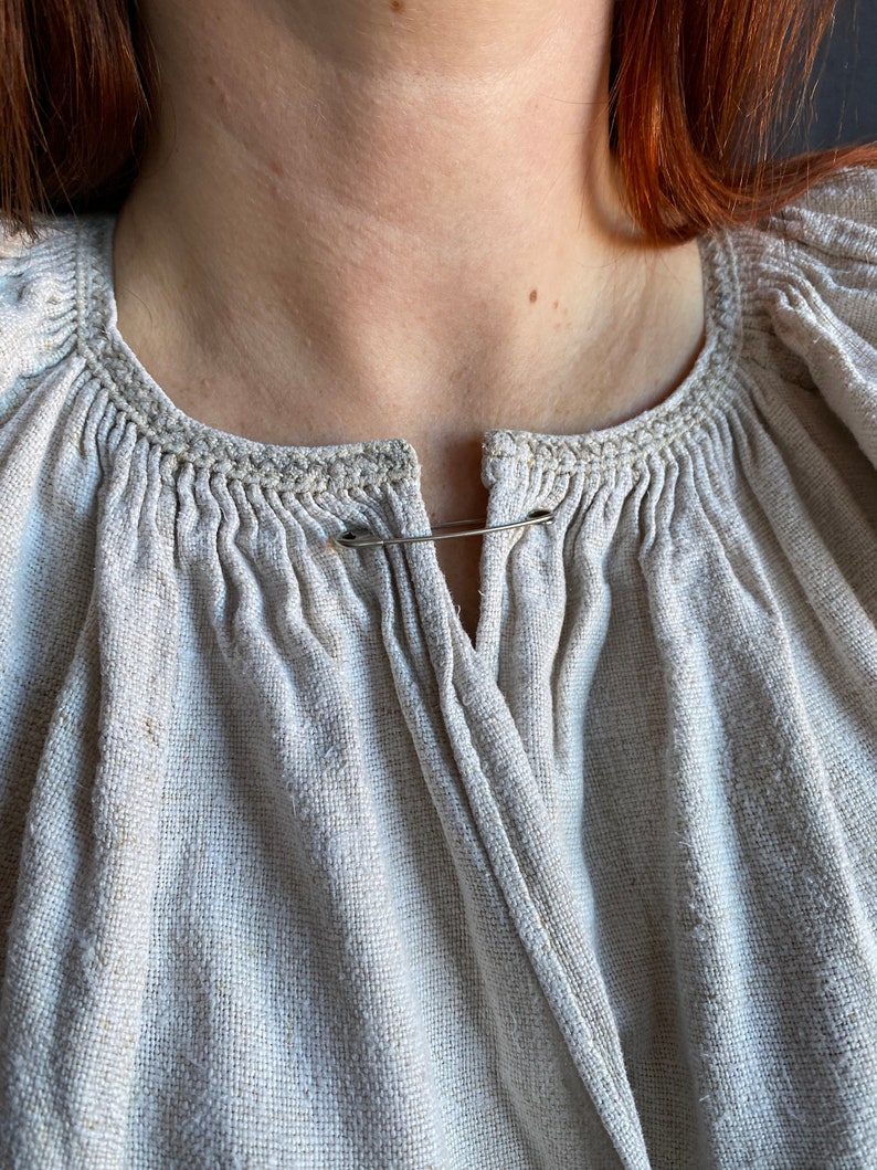 May include: A beige, textured linen blouse featuring a round neckline with gathered fabric details and a vertical slit opening at the centre. The garment is fastened at the top of the opening with a simple silver-toned metal safety pin.