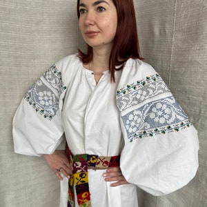 May include: A white embroidered blouse with long sleeves and a colorful woven belt. The blouse has a traditional Ukrainian design with floral patterns.
