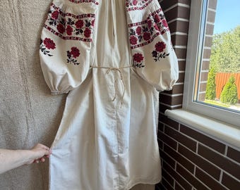May include: An off-white dress featuring voluminous sleeves with intricate red and black floral embroidery. The garment includes a simple tie-waist cord and a straight hemline, showcasing detailed needlework patterns across the upper sleeves.