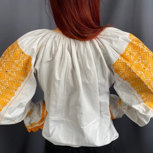 May include: White blouse with long sleeves and yellow embroidered details. The embroidery features a geometric pattern.