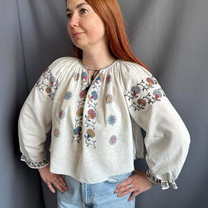 May include: A white linen blouse with embroidered floral designs in shades of blue, red, yellow, and brown. The blouse has a gathered neckline and long sleeves with embroidered cuffs.