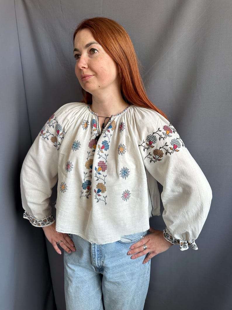 May include: A white linen blouse with embroidered floral designs in shades of blue, red, yellow, and brown. The blouse has a gathered neckline and long sleeves with embroidered cuffs.