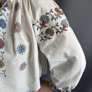 May include: A white linen blouse with colorful embroidered floral designs. The embroidery features a variety of colors, including red, blue, yellow, and green. The blouse has a gathered neckline and long sleeves.