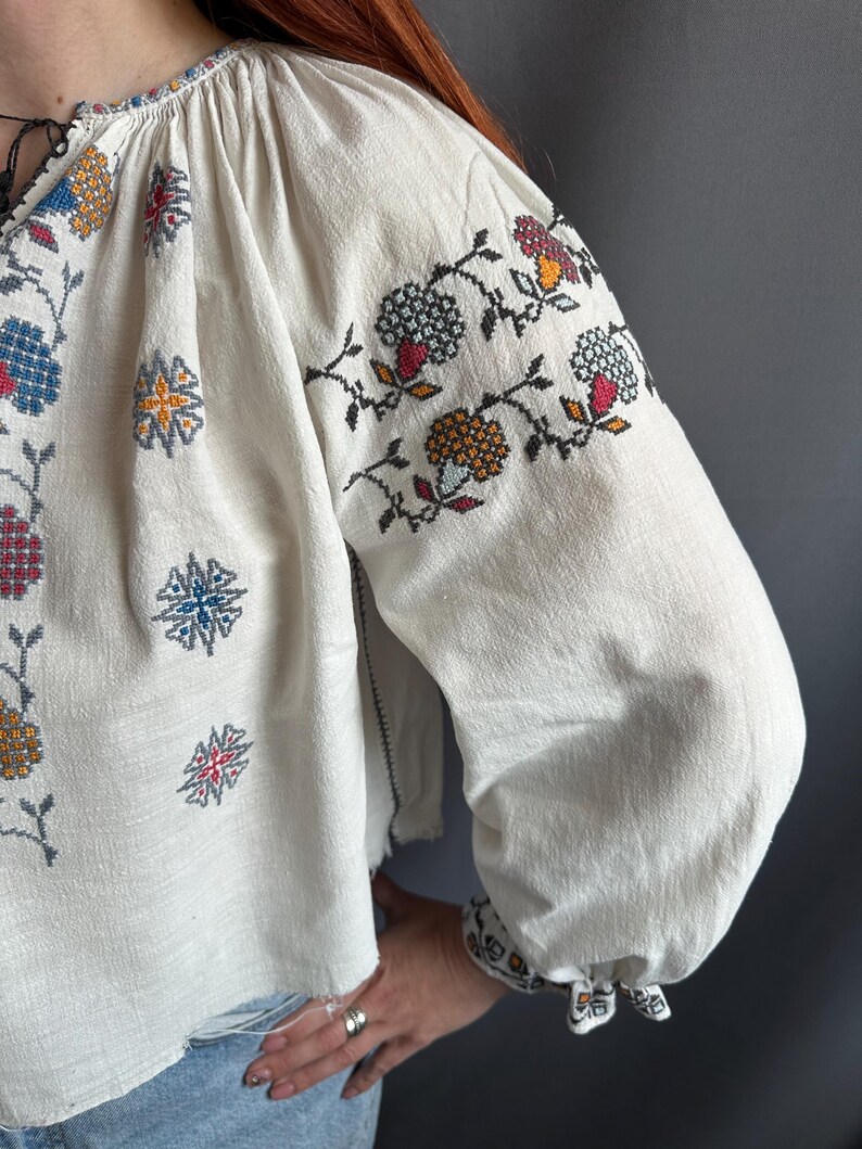 May include: A white linen blouse with colorful embroidered floral designs. The embroidery features a variety of colors, including red, blue, yellow, and green. The blouse has a gathered neckline and long sleeves.