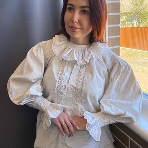 K&ouml;nnte beinhalten: Eine wei&szlig;e Bluse mit einem gro&szlig;en, gekr&auml;uselten Kragen und Spitzendetails. Die Bluse hat lange &Auml;rmel mit R&uuml;schen an den Manschetten.