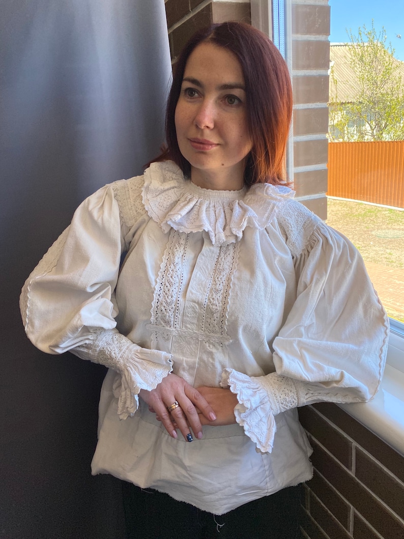 K&ouml;nnte beinhalten: Eine wei&szlig;e Bluse mit einem gro&szlig;en, gekr&auml;uselten Kragen und Spitzendetails. Die Bluse hat lange &Auml;rmel mit R&uuml;schen an den Manschetten.