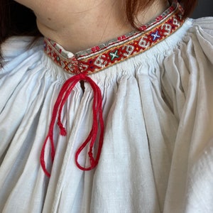 May include: White linen blouse with a red embroidered neckline and a red ribbon tied at the neck.