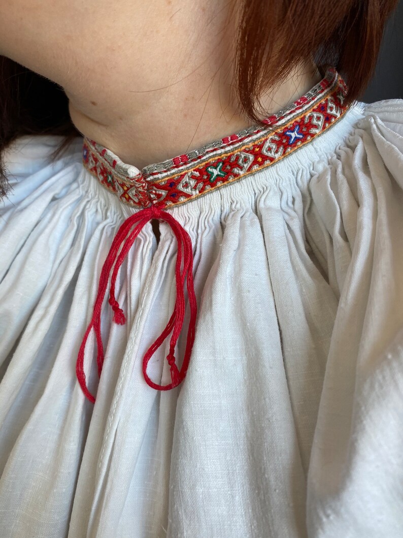 May include: White linen blouse with a red embroidered neckline and a red ribbon tied at the neck.