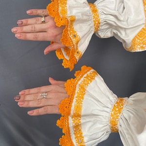 May include: White blouse with long sleeves and yellow embroidered details. The sleeves have a ruffled design and a crocheted trim.